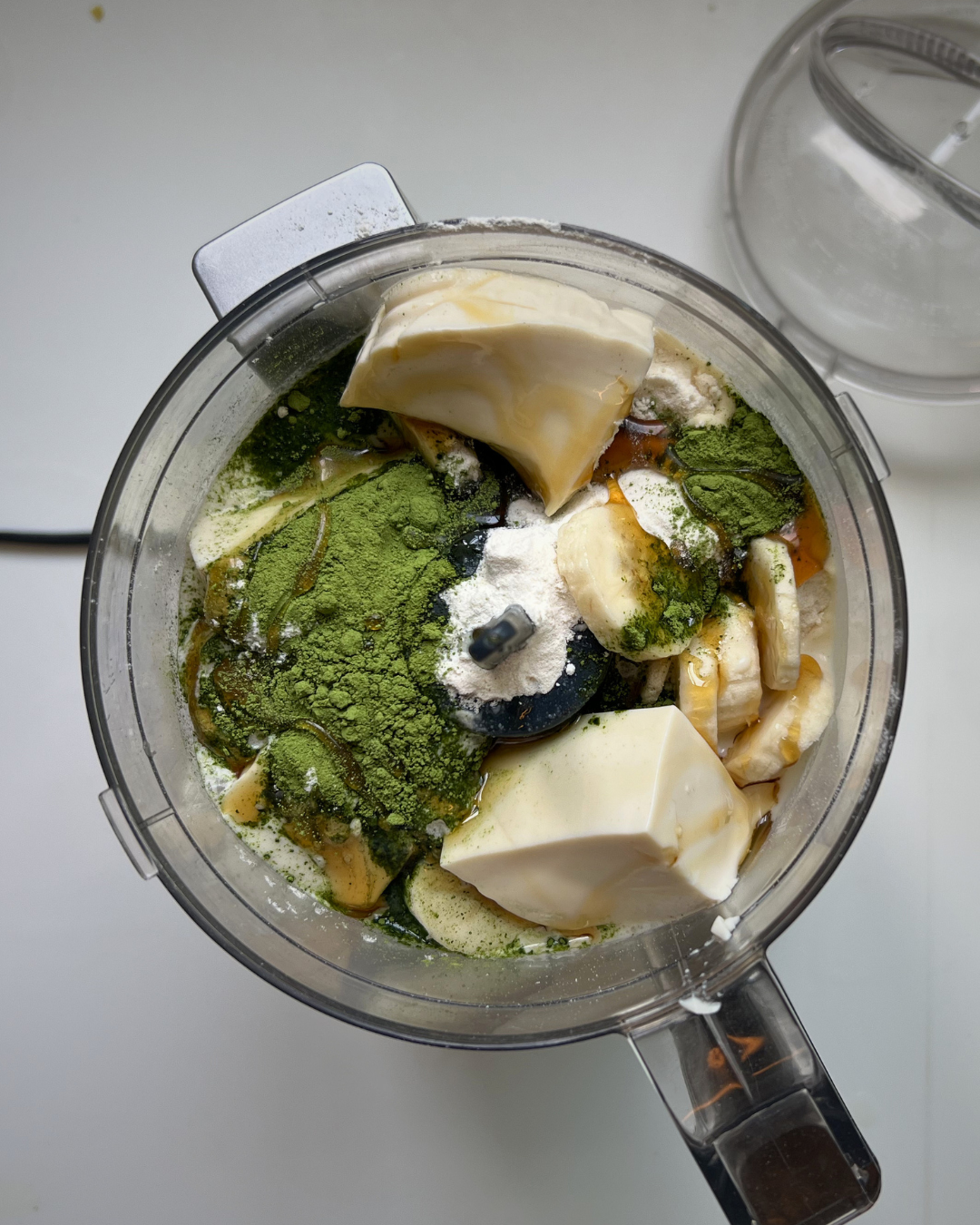 A top-down view of a food processor bowl filled with ingredients for a green smoothie, including banana slices, matcha green powder, white powder, and chunks of creamy white ingredient, with the clear plastic lid resting beside it on a white surface.