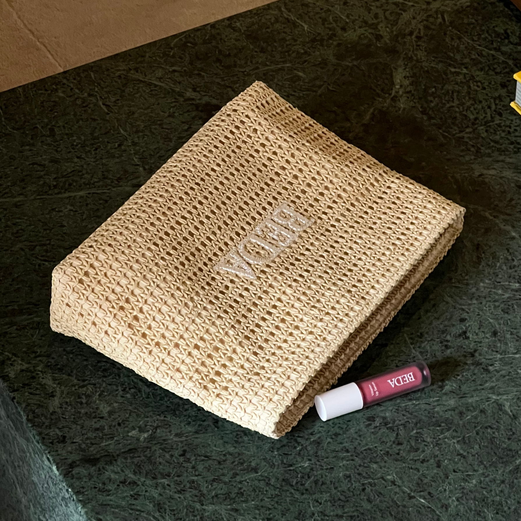 raffia travel pouch with a pink lipgloss on a green marble coffee table