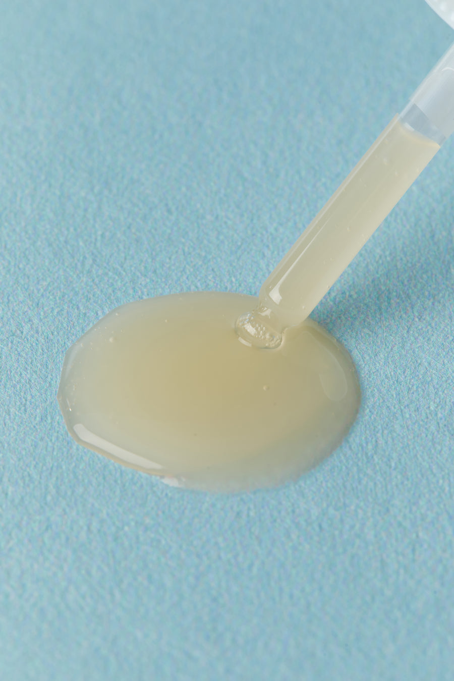 A close-up of a translucent pale yellow liquid gel or serum being dispensed from a clear pipette onto a textured light blue surface.