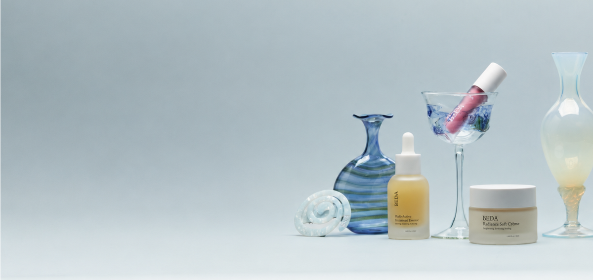 A styled product lineup on a light gray studio background featuring a pink lip gloss in a martini glass, a blue striped glass bottle, a clear textured glass object, a beige dropper bottle, and a white cream jar labeled REDA.
