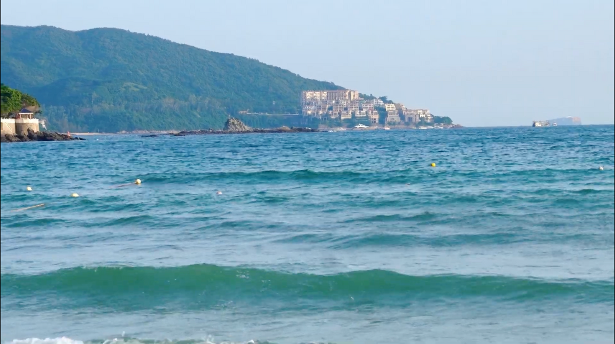 A scenic coastal view with turquoise ocean water in the foreground, a green hillside town in the distance, and a pale blue sky above.