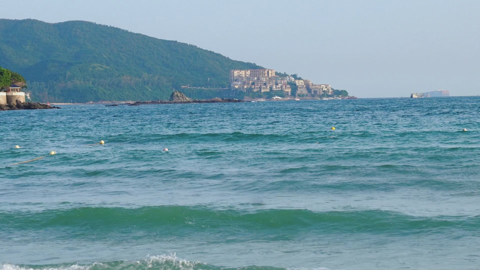 A scenic coastal view with turquoise ocean water in the foreground, a green hillside town in the distance, and a pale blue sky above.