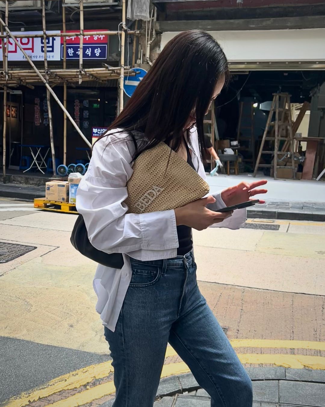 Woman walking on a street holding a woven raffia pouch with 'Beda' branding.