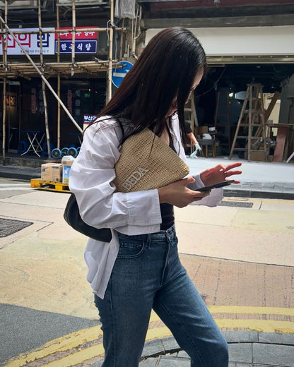 Woman walking on a street holding a woven raffia pouch with 'Beda' branding.